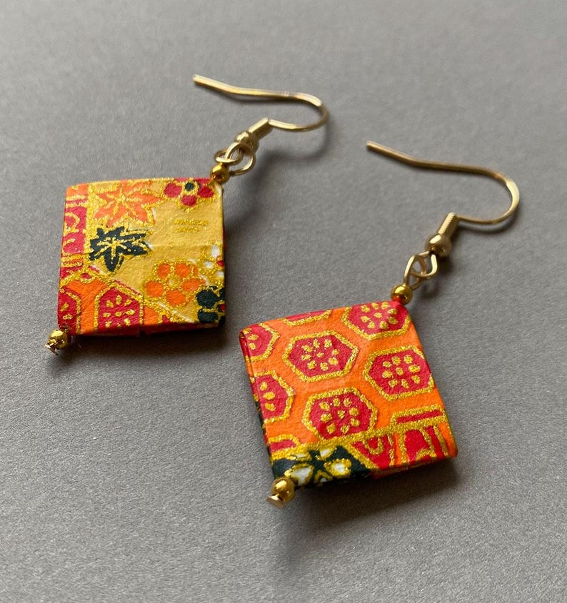K&ouml;nnte beinhalten: Ein Paar quadratische H&auml;ngeohrringe. Die Ohrringe haben goldfarbene Haken und quadratische Anh&auml;nger mit einem farbenfrohen, gemusterten Design in Orange, Rot und Gold. Jeder Ohrring hat eine kleine goldene Perle unten.
