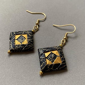 K&ouml;nnte beinhalten: Handgefertigte Ohrringe mit quadratischem Design, goldenen und schwarzen Akzenten. Die Ohrringe haben goldfarbene Haken und kleine goldene Perlen unten. Die schwarzen Bereiche sind mit kleinen goldenen Punkten versehen.