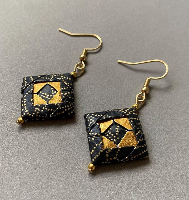 K&ouml;nnte beinhalten: Handgefertigte Ohrringe mit quadratischem Design, goldenen und schwarzen Akzenten. Die Ohrringe haben goldfarbene Haken und kleine goldene Perlen unten. Die schwarzen Bereiche sind mit kleinen goldenen Punkten versehen.