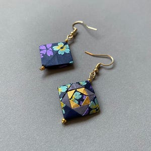 K&ouml;nnte beinhalten: Ein Paar Origami-Ohrringe mit quadratischer Form. Die Ohrringe haben ein dunkelblaues Blumenmuster mit lila und gelben Blumen. Sie haben goldfarbene Ohrhaken und kleine goldene Perlen unten.