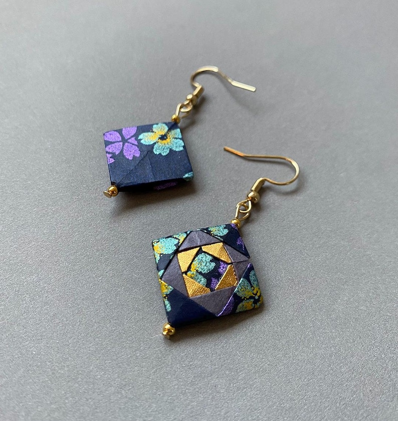 K&ouml;nnte beinhalten: Ein Paar Origami-Ohrringe mit quadratischer Form. Die Ohrringe haben ein dunkelblaues Blumenmuster mit lila und gelben Blumen. Sie haben goldfarbene Ohrhaken und kleine goldene Perlen unten.