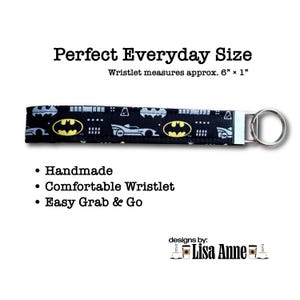 Może przedstawiać: Czarna brelok-opaska na nadgarstek z tkaniny z powtarzającym się wzorem ż&oacute;łtych logo Batmana i szarych sylwetek samochod&oacute;w. Opaska zawiera srebrne metalowe zapięcie i okrągłe k&oacute;łko na klucze. Tekst na obrazku: Perfect Everyday Size, Wristlet measures approx. 6&rdquo; x 1&rdquo;, Handmade, Comfortable Wristlet, Easy Grab & Go, designs by: Lisa Anne.