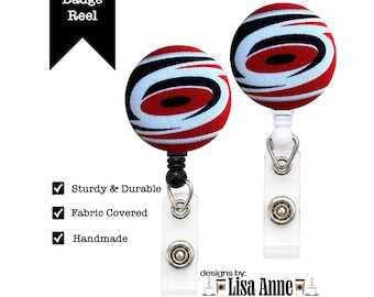 May include: Two round badge reels featuring a fabric-covered top with a red, black, and white swirling graphic pattern. Each reel has a clear plastic strap with a metal snap button. Text includes Badge Reel, Sturdy & Durable, Fabric Covered, Handmade, and designs by: Lisa Anne.