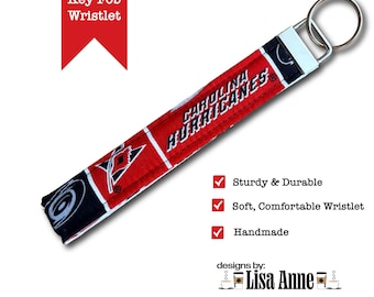 May include: A fabric key fob wristlet featuring the Carolina Hurricanes logo and team name in red, black, and white. The strap is attached to a silver-toned metal hardware clamp and a circular split ring. Text includes Key Fob Wristlet and designs by: Lisa Anne.