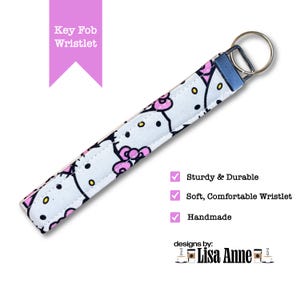 May include: A fabric key fob wristlet featuring a repeating Hello Kitty pattern on a light blue background. The wristlet includes a dark blue metal hardware clamp and a silver-toned circular split ring. Text on the image reads Key Fob Wristlet, Sturdy & Durable, Soft, Comfortable Wristlet, Handmade, and designs by: Lisa Anne.