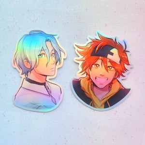 Puede incluir: Dos pegatinas holográficas con personajes de estilo anime. El personaje de la izquierda tiene el pelo azul y lleva una camisa blanca. El personaje de la derecha tiene el pelo naranja y lleva una sudadera amarilla.