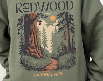 May include: Olive green sweatshirt with a Redwood National Park graphic. The design features a path through a forest with large trees, a sun, and the text "REDWOOD NATIONAL PARK".