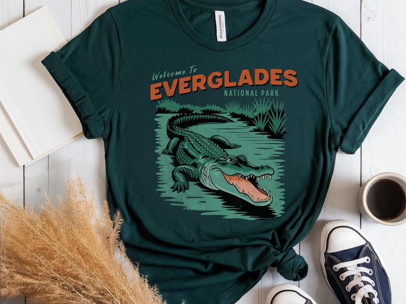 May include: Dark green t-shirt with a graphic of an alligator in a swamp, and the text "Welcome to Everglades National Park." The design uses green, orange, and black. The shirt is styled with blue trainers and a coffee cup.