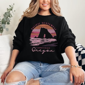 Peut inclure: Un sweat-shirt noir avec un motif graphique rose et orange d'un coucher de soleil sur une formation rocheuse. Le graphique indique "Pacific Northwest Cannon Beach Oregon".