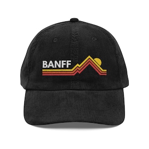 May include: Black corduroy baseball cap with the word "BANFF" in white block letters. The cap features a mountain and sun graphic in yellow, orange, and red. The hat is a classic six-panel design with a curved brim.