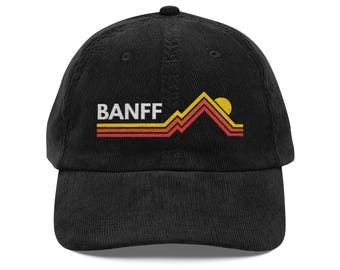 May include: Black corduroy baseball cap with the word "BANFF" in white block letters. The cap features a mountain and sun graphic in yellow, orange, and red. The hat is a classic six-panel design with a curved brim.
