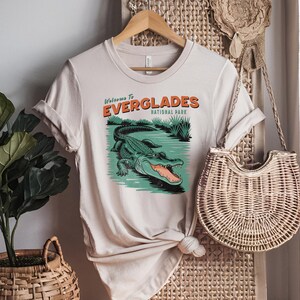 May include: Beige t-shirt featuring an alligator graphic and the text "Welcome to Everglades National Park". The design is in orange and green. The t-shirt is displayed on a wooden hanger.