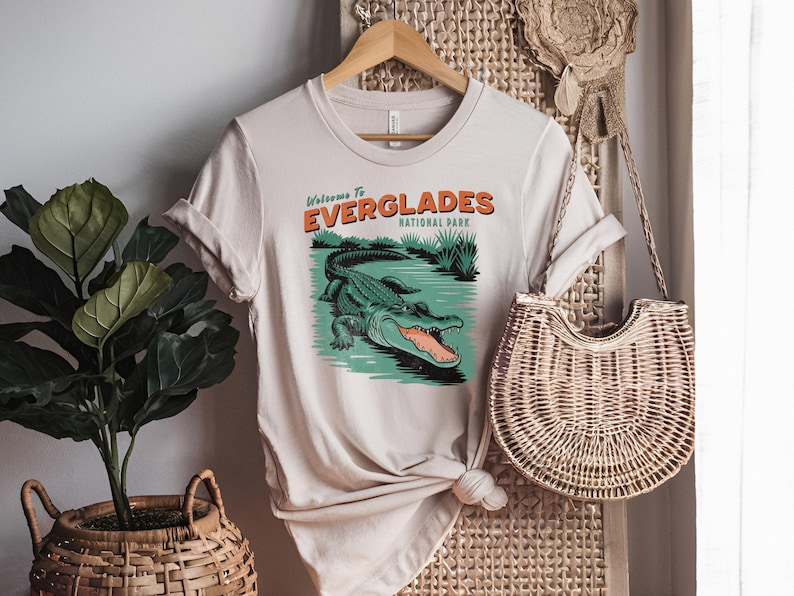 May include: Beige t-shirt featuring an alligator graphic and the text "Welcome to Everglades National Park". The design is in orange and green. The t-shirt is displayed on a wooden hanger.