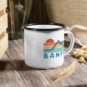 May include: White enamel mug with a black rim and handle. The mug features a retro-style graphic of mountains and a sunset with the word "BANFF" in blue text.