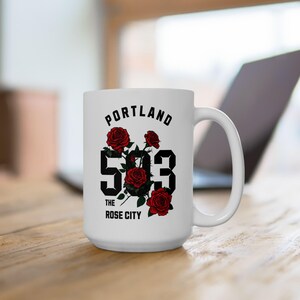 Könnte beinhalten: Eine weiße Keramiktasse mit einem schwarzen und roten Design. Die Tasse zeigt den Text "PORTLAND THE ROSE CITY" und die Nummer "503" mit roten Rosen, die die Zahlen umgeben.