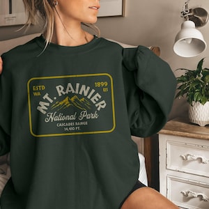 Puede incluir: Una sudadera de color verde oscuro con un gráfico de estilo vintage del Parque Nacional del Monte Rainier. El gráfico presenta el texto "Mt. Rainier National Park" con las palabras "Estd WA 1899 US" y "Cascades Range 14,410 FT" en texto más pequeño.