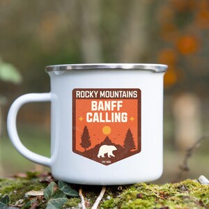 May include: White enamel camping mug with a brown and orange sticker that says "Rocky Mountains Banff Calling" with a silhouette of a bear and trees. The sticker also says "Est. 1885".