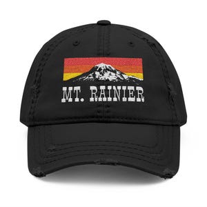 May include: Black distressed baseball cap featuring a rectangular graphic of Mount Rainier with a red, orange, and yellow sunset. The text "MT. RAINIER" is printed in white below the graphic. The cap has a curved brim and a distressed finish.