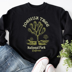 Pode incluir: Camisola de moletom preta com um gráfico verde do logotipo do Parque Nacional Joshua Tree. O logotipo apresenta uma árvore de Josué com o texto "Joshua Tree National Park California".