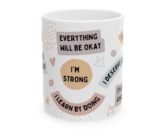 Pode incluir: Caneca de cerâmica branca decorada com frases inspiradoras como "Tudo ficará bem" e "Eu sou forte". O design inclui formas e texto em cores pastel, criando uma estética positiva e edificante.