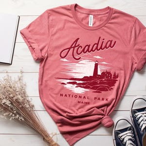 May include: A pink t-shirt with a red graphic of a lighthouse and the text "Acadia National Park Maine".
