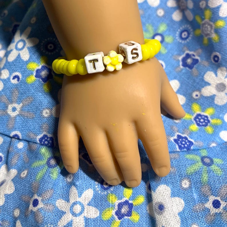 May include: A yellow and white beaded bracelet with the letters "T.S." and a yellow flower charm.
