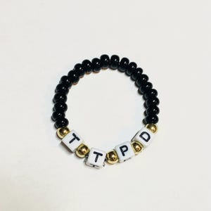 May include: Black beaded bracelet with white letter beads spelling out "TPD".