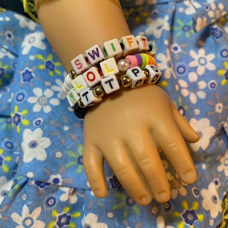May include: A child's wrist wearing a stack of colorful beaded bracelets with the words "SWIFT", "LOL", "T", and "PD" spelled out.