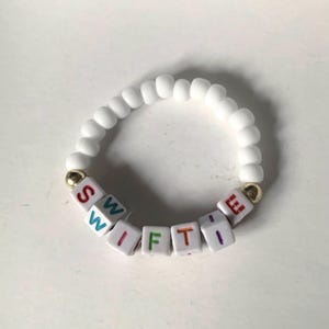May include: White beaded bracelet with the word "SWIFTIE" spelled out in colorful letter beads.