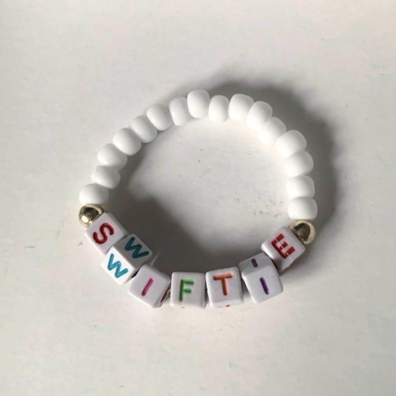 May include: White beaded bracelet with the word "SWIFTIE" spelled out in colorful letter beads.