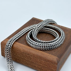 Op de afbeelding: Een zilveren ketting met een geweven ontwerp. De ketting is opgerold op een houten oppervlak.