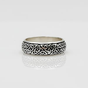 May include: A silver ring with a detailed Celtic knot pattern. The ring has a polished finish and a wide band. The intricate design covers the entire outer surface of the ring, creating a textured appearance. The ring is displayed on a white surface.