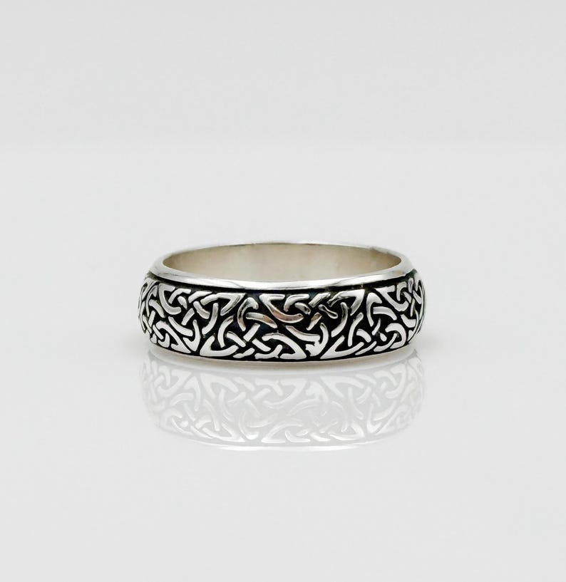 May include: A silver ring with a detailed Celtic knot pattern. The ring has a polished finish and a wide band. The intricate design covers the entire outer surface of the ring, creating a textured appearance. The ring is displayed on a white surface.