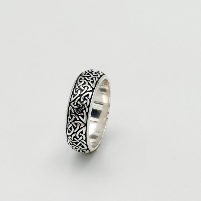 May include: A silver ring with a Celtic knot design. The ring features an intricate pattern of interwoven lines and shapes, creating a continuous, symmetrical design. The ring is made of a shiny, reflective metal, and the design is in a darker tone.