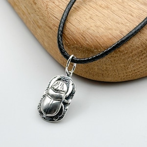 May include: A silver scarab beetle pendant on a black leather cord. The pendant is detailed with a textured surface and the beetle is depicted in a realistic style.