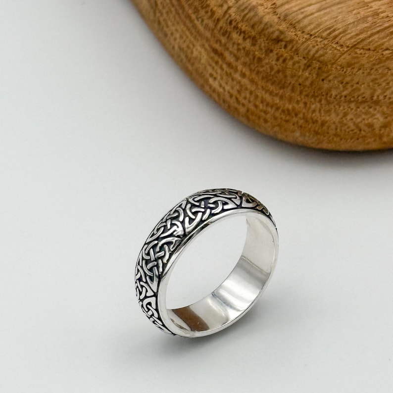 May include: A silver ring with a Celtic knot design. The ring has a polished finish and a detailed pattern of interwoven lines. The ring is displayed on a white surface with a wooden element in the background.