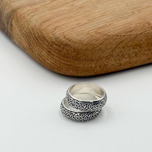 May include: Two silver rings with a Celtic knot design are stacked on a white surface. The rings have a detailed pattern around the band. A wooden cutting board is in the background.