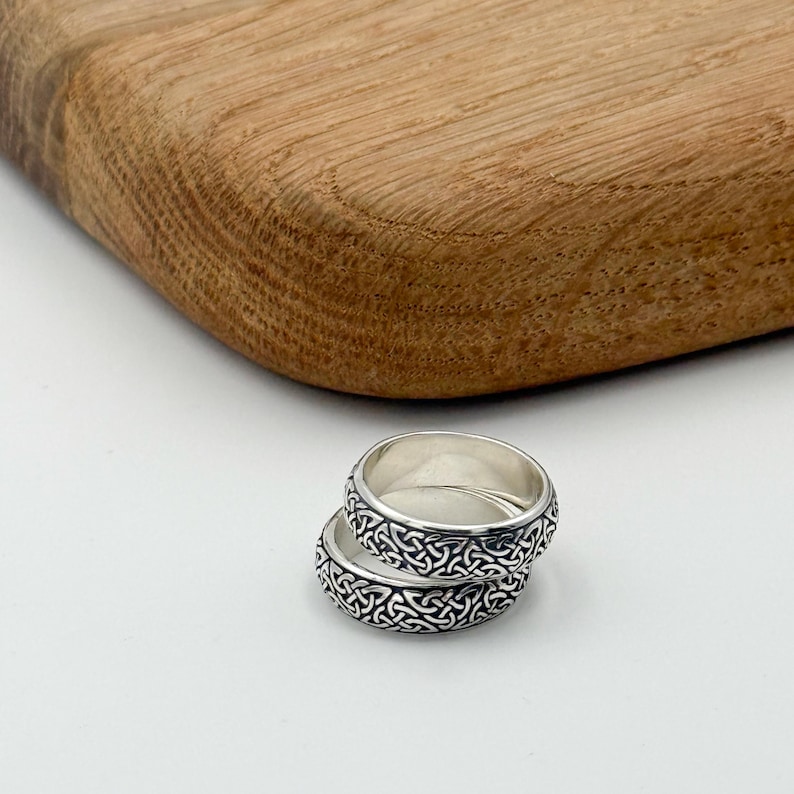 May include: Two silver rings with a Celtic knot design are stacked on a white surface. The rings have a detailed pattern around the band. A wooden cutting board is in the background.