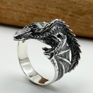 Könnte beinhalten: Ein detaillierter silberner Drachenring. Der Kopf und Körper des Drachen umschließen das Ringband, mit strukturierten Schuppen und Flügeldarstellungen. Der Ring besteht aus glänzendem Metall.