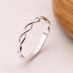 Könnte beinhalten: Ein silberner Ring mit geflochtenem Design. Der Ring hat eine polierte Oberfläche und einen schlichten, eleganten Stil. Das verschlungene Muster erzeugt eine durchgehende Schleife und zeigt eine klassische und zeitlose Ästhetik.