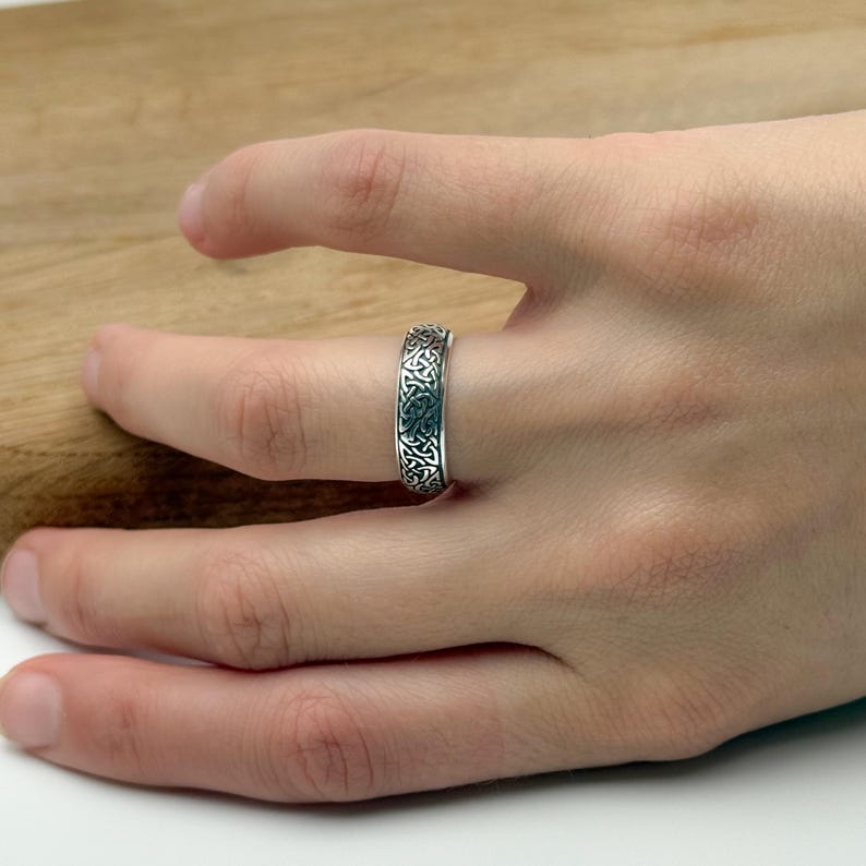 May include: A silver ring with an intricate Celtic knot design. The ring is worn on a finger, showcasing the detailed pattern of interwoven lines and shapes. The ring's surface reflects light, highlighting its metallic sheen.