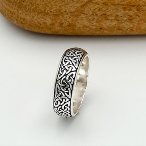 May include: A silver ring with a Celtic knot design. The ring has a polished finish and features an intricate pattern of interwoven lines. The ring is a band style, with the design wrapping around the entire circumference.