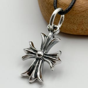 May include: A silver pendant with a decorative cross design. The cross has detailed, pointed edges and a central sphere. It hangs from a silver loop attached to a black cord, a classic piece of jewellery.
