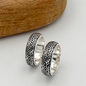 May include: Two silver rings with a Celtic knot design. The rings have a polished finish and a detailed, interwoven pattern in black against the silver. The rings are displayed on a white surface with a wooden element in the background.