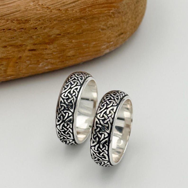 May include: Two silver rings with a Celtic knot design. The rings have a polished finish and a detailed, interwoven pattern in black against the silver. The rings are displayed on a white surface with a wooden element in the background.