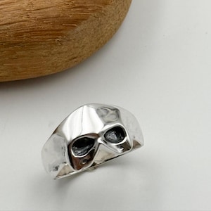 Könnte beinhalten: Ein silberner Totenkopfring mit dunklen Augenhöhlen. Der Ring hat eine polierte Oberfläche und ein detailliertes Totenkopfdesign. Der Ring liegt auf einer weißen Oberfläche mit einem Holzobjekt im Hintergrund.