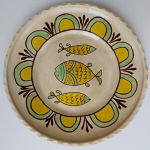 May include: A cream-colored ceramic plate with a scalloped edge. The plate features a central design of three yellow and green fish. A border of green and yellow arches and brown accents surrounds the fish.