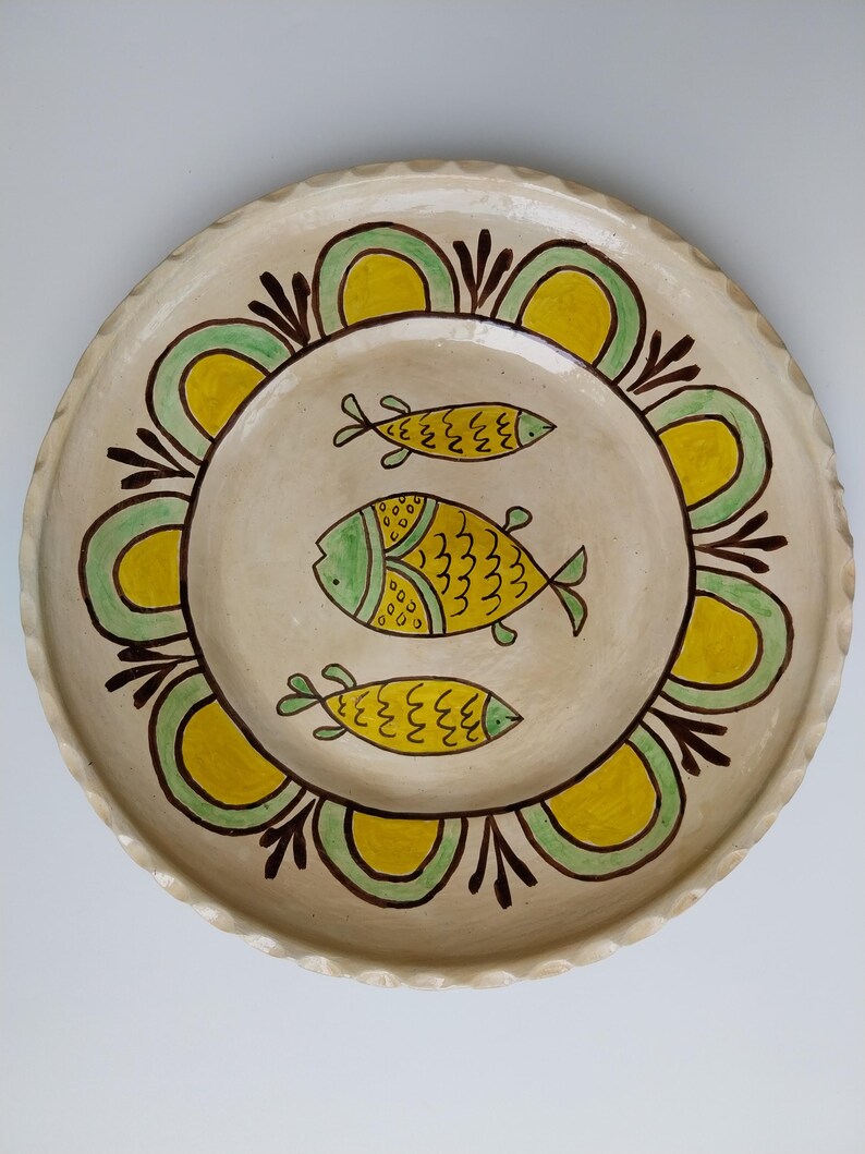 May include: A cream-colored ceramic plate with a scalloped edge. The plate features a central design of three yellow and green fish. A border of green and yellow arches and brown accents surrounds the fish.