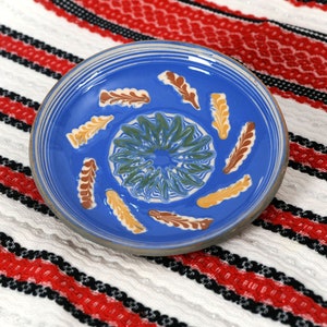 May include: A blue ceramic plate with a white and green floral design and yellow and brown accents. The plate is on a white and red striped fabric.