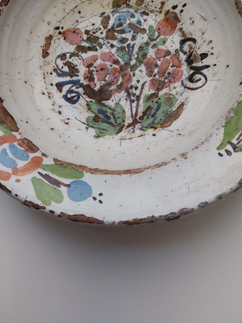 May include: A white ceramic plate featuring a central floral design. The flowers are painted in pink, orange, and blue, with green foliage. The plate's rim is a dark brown, and the overall aesthetic suggests a rustic, aged finish.
